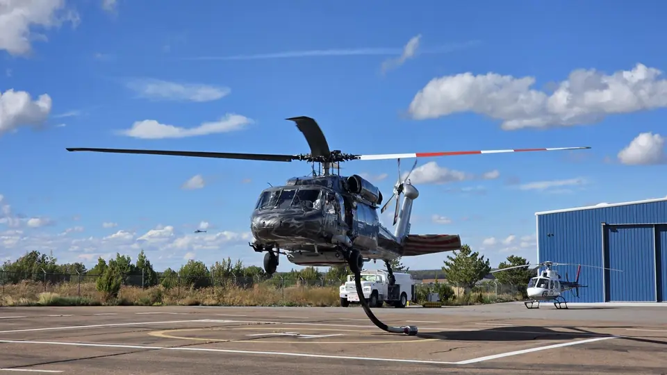 UH-60 Black Hawk in a custom flag livery lifting off a helipad beside a blue hangar