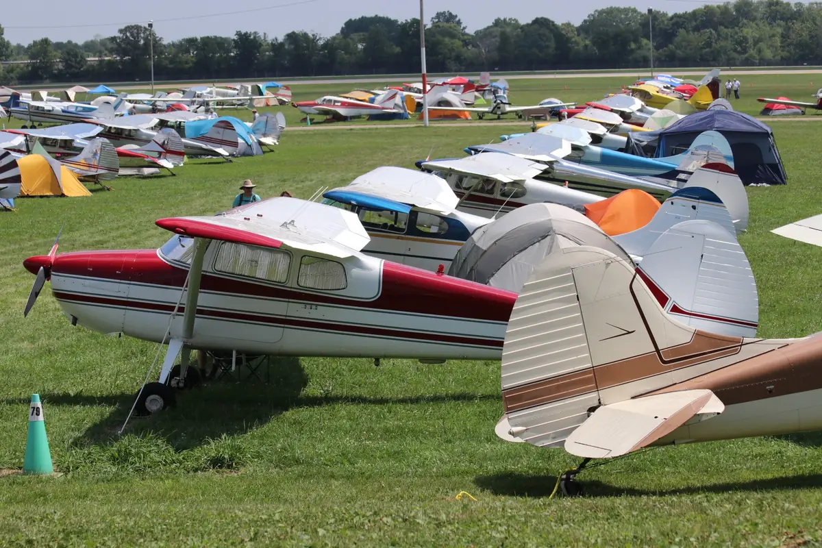 Classic high-wing airplanes parked in rows with camping tents beside them on the grass at Oshkosh