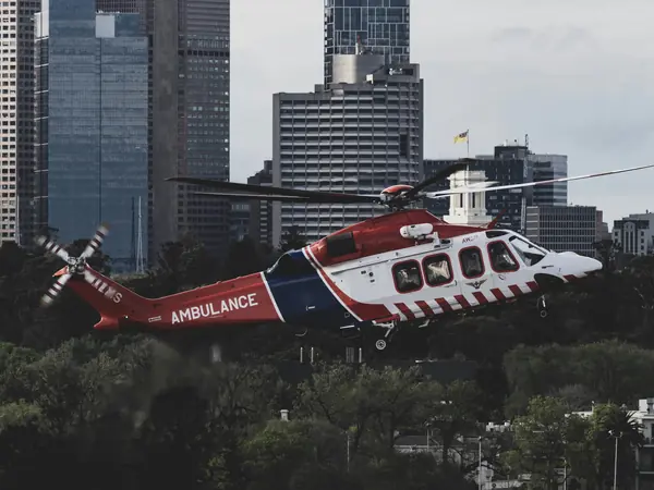 Red and white air ambulance helicopter flying low over trees, with city towers behind