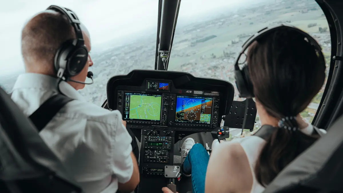 Pilot and passenger in a helicopter with Garmin glass-cockpit displays, flying over farmland